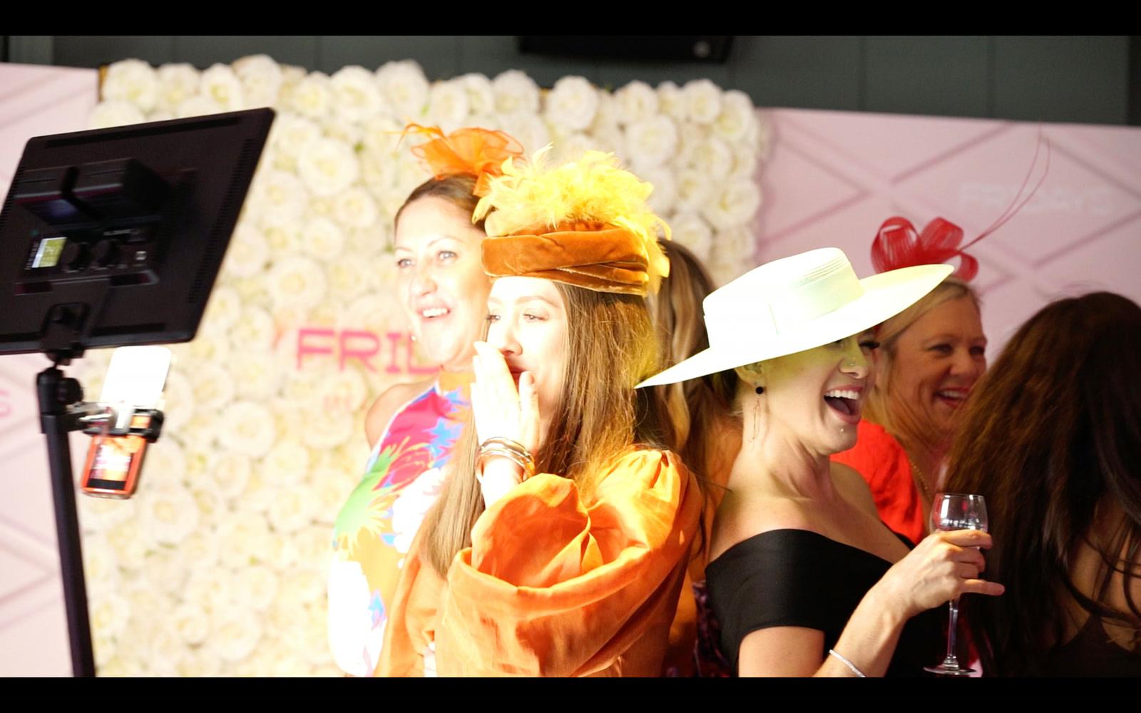 Guests in race-day hats laughing in front of a flower-wall photo booth backdrop at an event